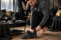 Homme enfilant ses pantoufles fourrées gris charbon sur un plancher de bois froid un matin d'hiver, intérieur douillet et tasse de café sur la table basse
