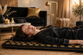 Femme détendue allongée sur le tapis d'acupression noir aux picots dorés DULUXE, moment de relaxation à la maison